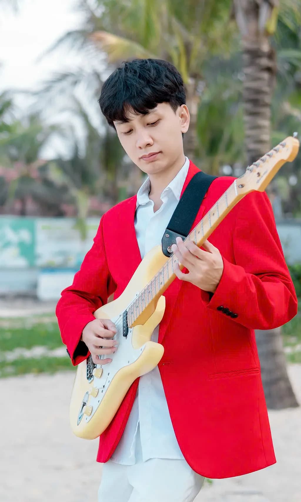 Guitarist in a red blazer playing a cream electric guitar on a palm-lined beach