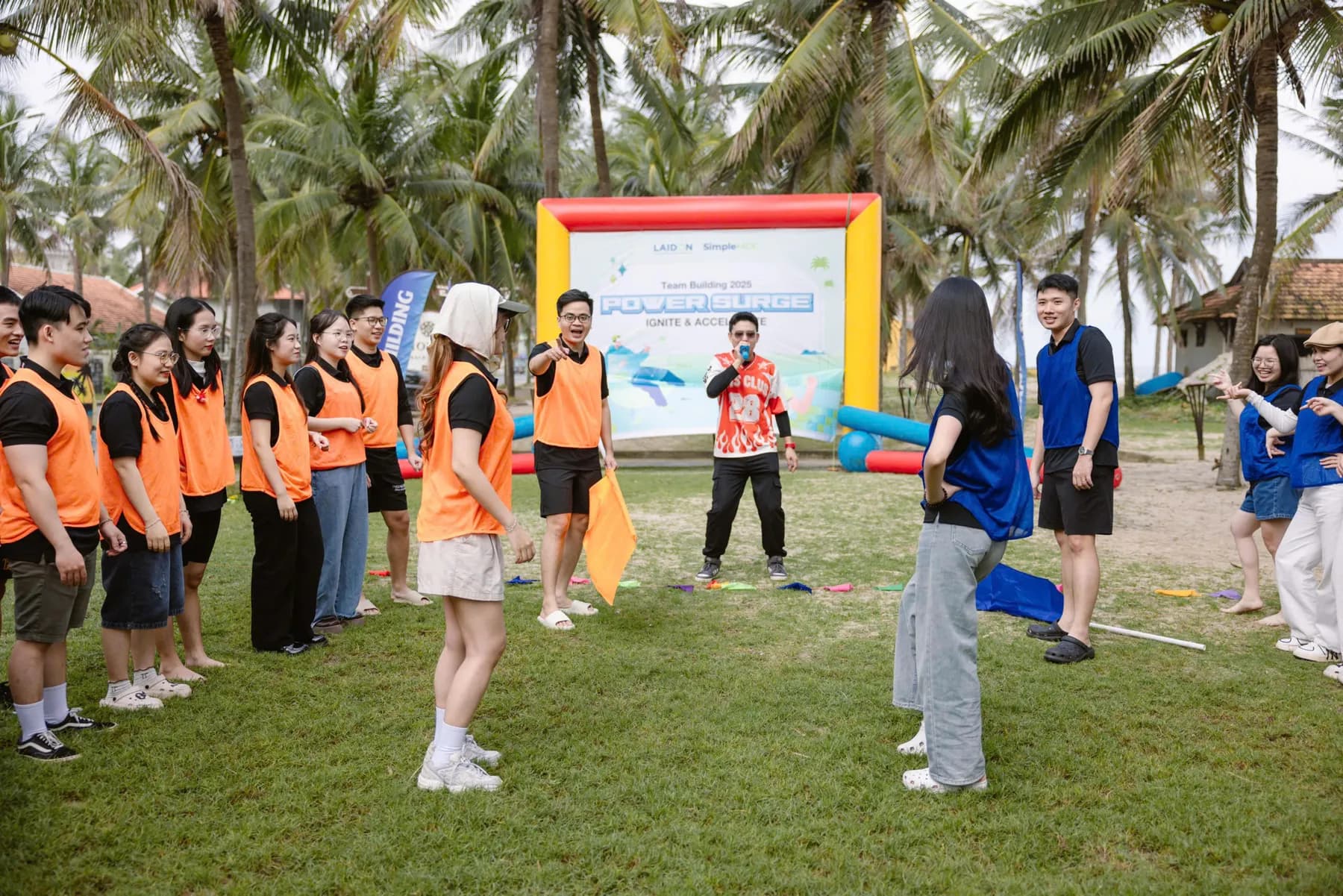 Two teams in orange and blue bibs facing off in a beach team building game led by an MC