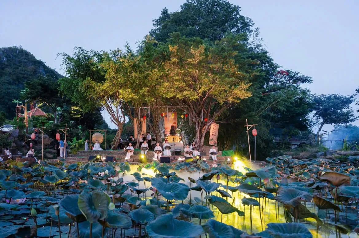Sound healing performers in white on a lantern-lit stage under a banyan tree, beyond a lotus pond at dusk