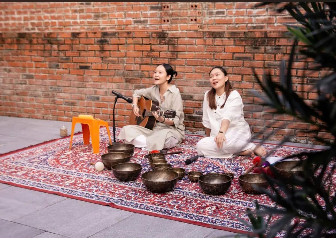 Two musicians with guitar and singing bowls on a patterned rug before a brick wall